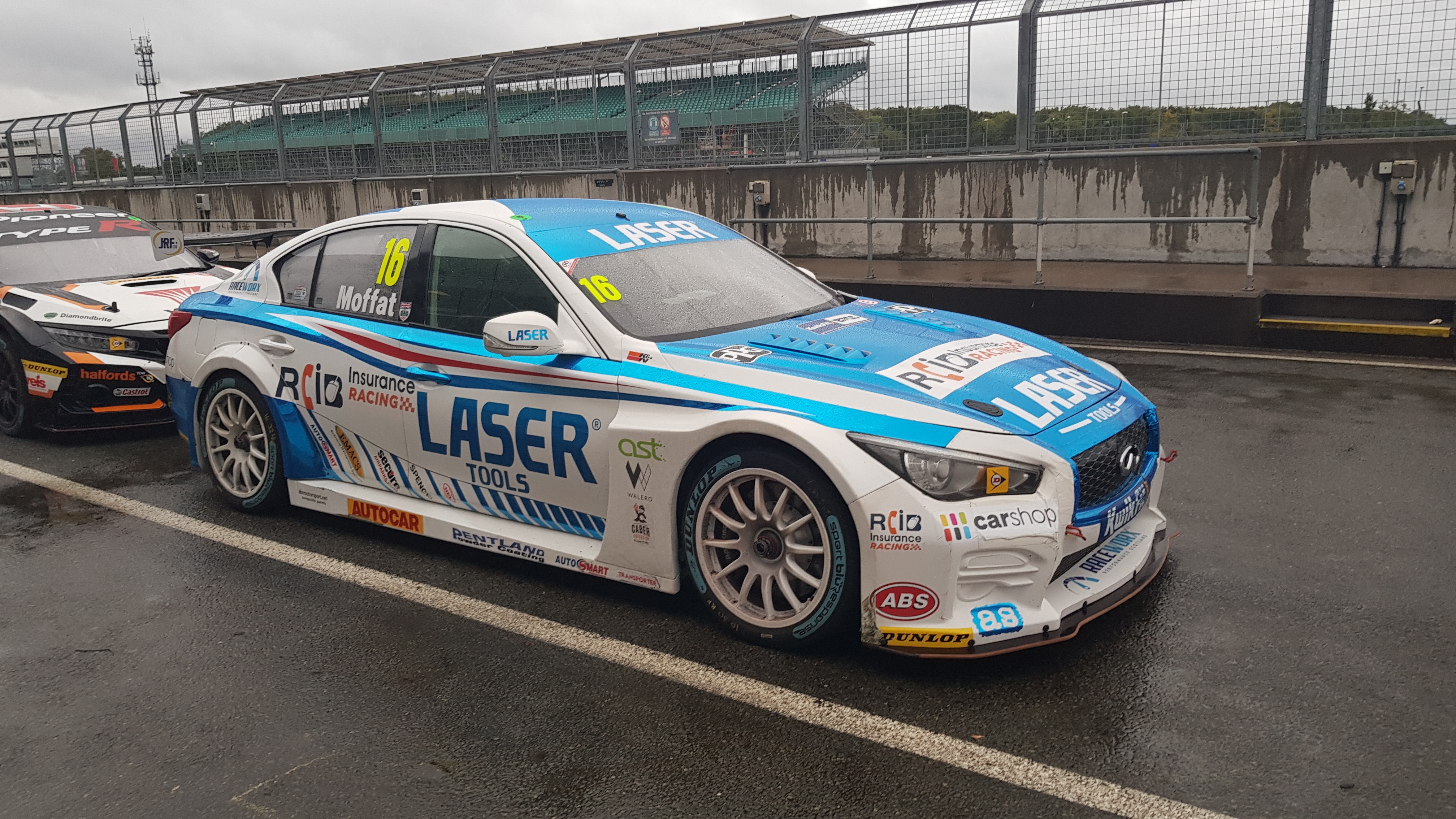 Front 3/4 photo of a white and light blue Infiniti Q50 in the pitlane at a damp Silverstone after a BTCC round